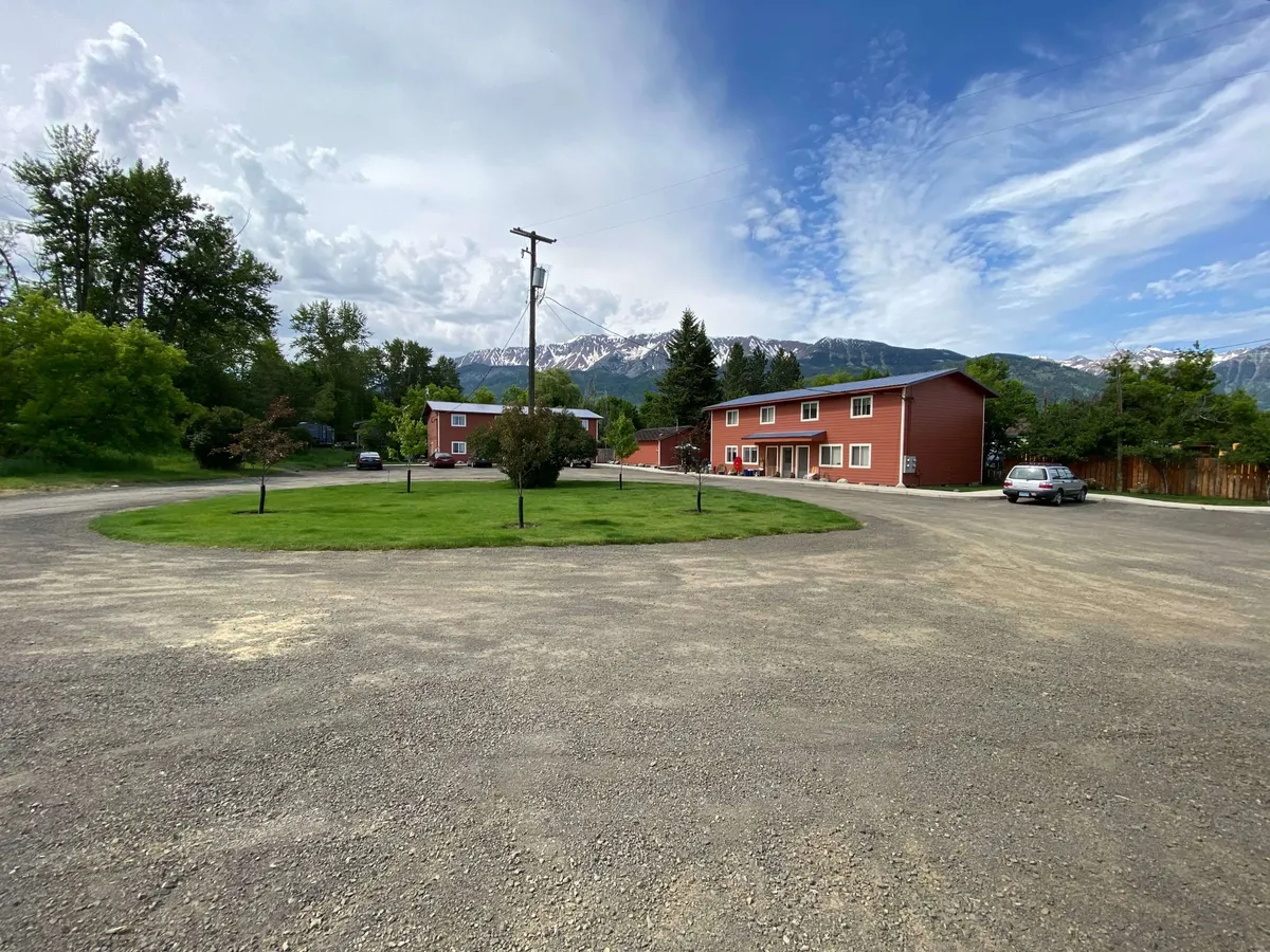 This exterior image showcases the condo's distinctive red buildings set against a breathtaking mountain backdrop, with a green lawn in the foreground. The scene captures a sense of peacefulness and provides a direct connection between the home and the outdoor environment. The setting encourages outdoor activities like strolling or picnicking while highlighting the mountain views that define the area.