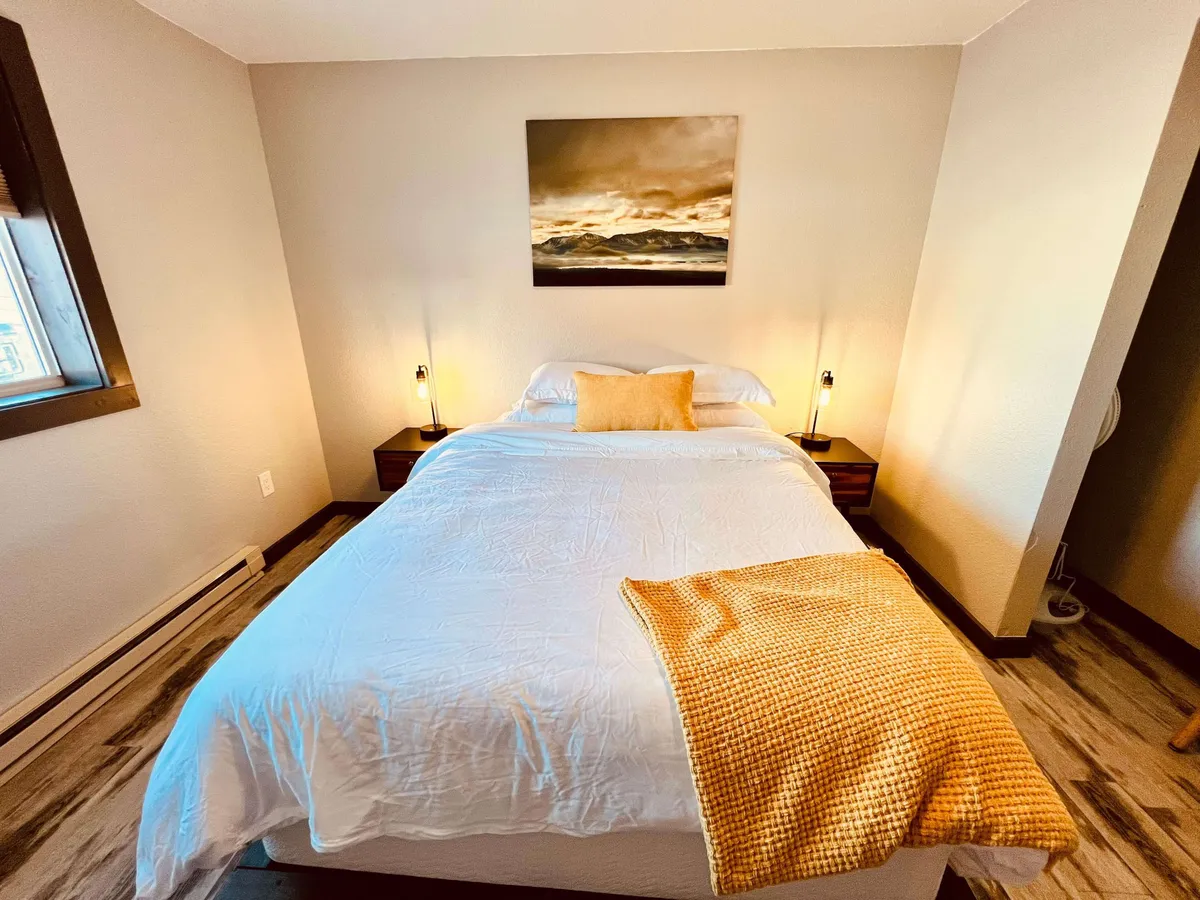 A tranquil bedroom featuring a plush bed adorned with a warm throw blanket, flanked by nightstands and subtle artwork. The soft glow of natural light enhances the serene, cozy ambiance, inviting guests to relax and recharge.
