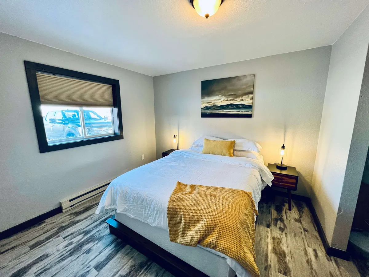 A bedroom centered around a cozy bed with soft, inviting linens. Framed artwork hangs above the headboard, and matching nightstands with lamps flank each side. Soft morning light filters through a window, creating a calm, restful atmosphere ideal for relaxing or reading.