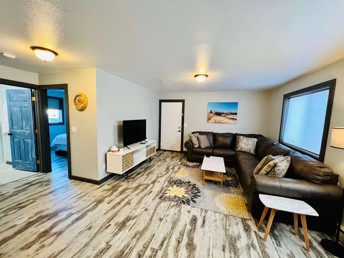 A cozy living room featuring a large leather sectional sofa, a coffee table, a television mounted on the wall, decorative wall art, and a plush area rug. The space is bathed in natural light from nearby windows, creating an inviting atmosphere ideal for socializing, relaxing, or watching TV.