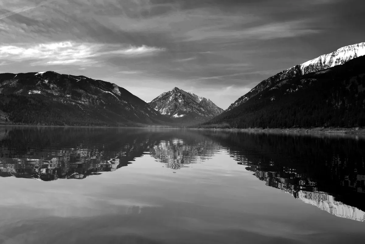 A wide view of a calm lake bordered by a striking mountain range under a cloudy sky. The serene water and natural setting evoke opportunities for hiking, wildlife observation, and outdoor adventure nearby.